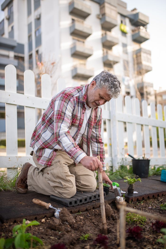 A senior man kneeling and gardening with tools in an urban outdoor setting.